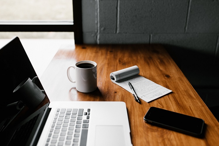 Wooden desk with laptop, coffee mug, smartphone and open notepad in white cinderblock room