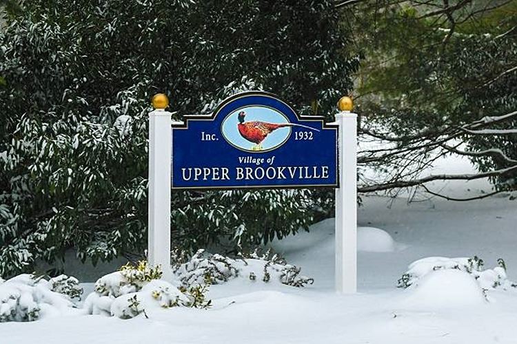 Sign designating entrance to the Village of Upper Brookville, New York amid snow and pine trees