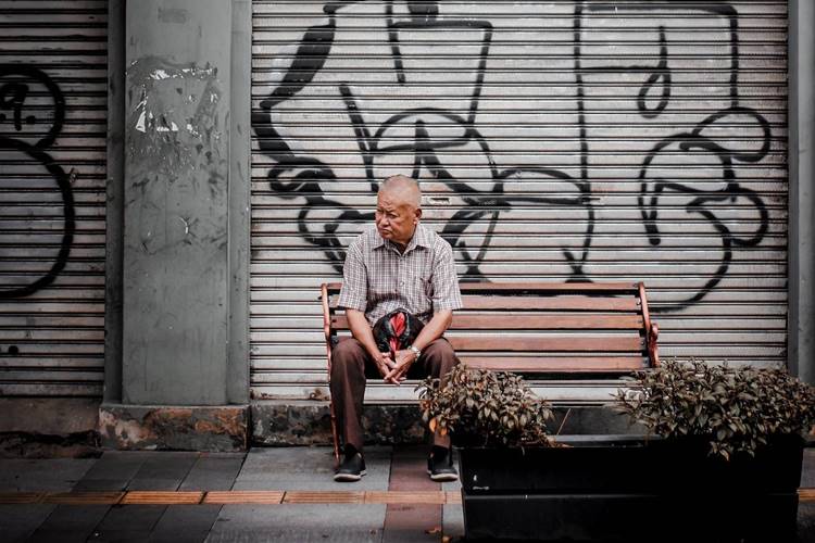 Elderly man in short-sleeved dress shirt on park bench beside steel rolling garage door with black graffiti