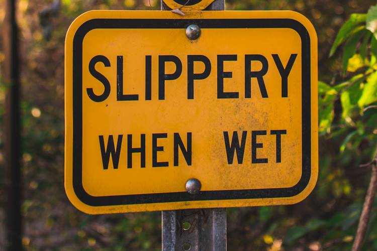 Yellow road sign reading 'SLIPPERY WHEN WET' near sunlit trees and telephone pole
